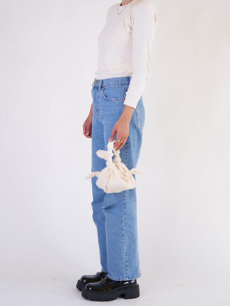 Person wearing blue jeans and a white top, holding a small white shaggy chiffon knotted bag on a plain background
