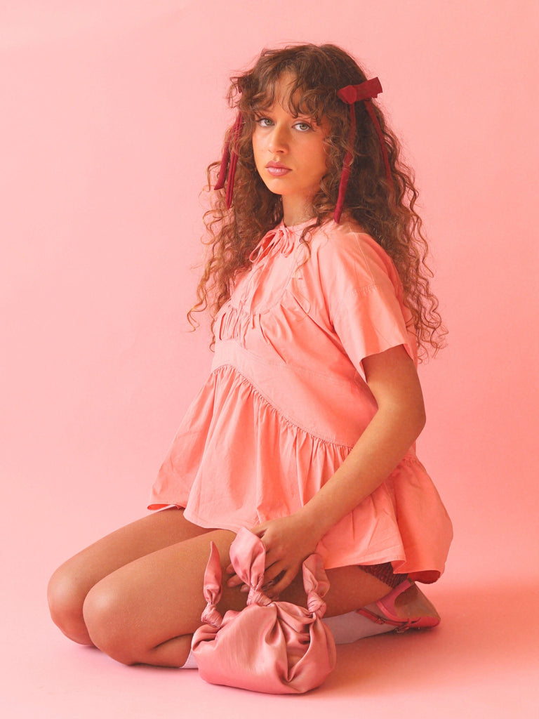 Woman in a pink outfit with two burgundy bow hair clips and a pink knotted handbag on a pink background