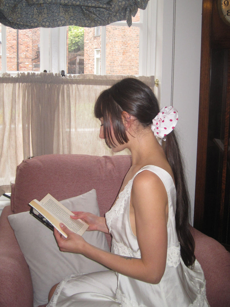 Woman in a white dress sitting on a couch reading a book with a window and curtains in the background. She wears a white and pink polka dot scrunchie in her ponytail