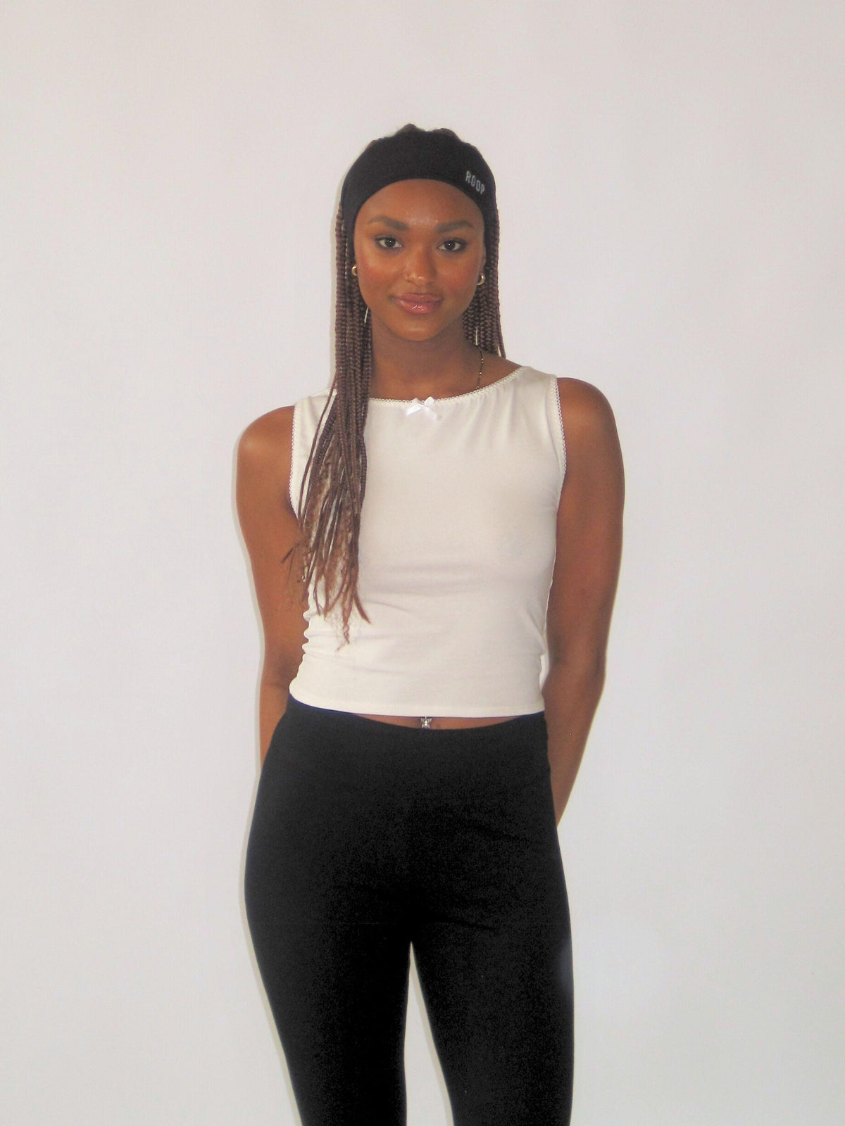 Woman wearing a white sleeveless top and black pants against a plain background. She wears a black stretch headband.