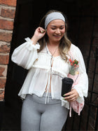 Woman holding a coffee cup and flowers against a brick wall. She wears a white flowy top and a grey stretch headband