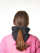 Person wearing a large black bow  scrunchie in their brown hair against a white background. They are wearing a pink knit cardigan.