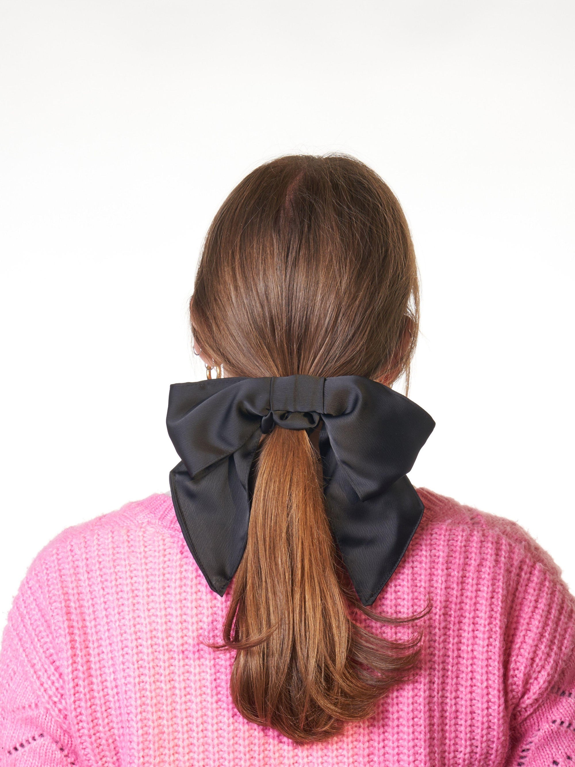 Person wearing a large black bow  scrunchie in their brown hair against a white background. They are wearing a pink knit cardigan.