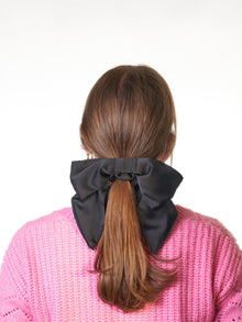 Person wearing a large black bow  scrunchie in their brown hair against a white background. They are wearing a pink knit cardigan.