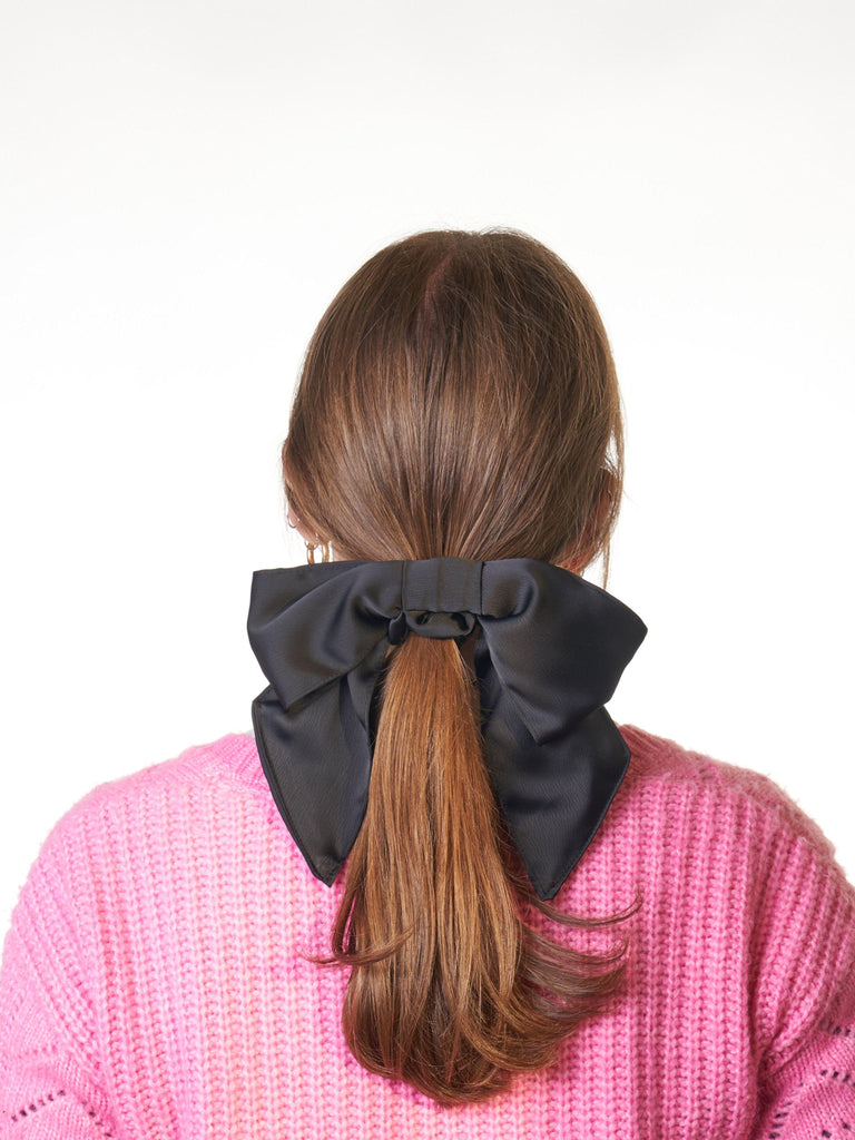 Person wearing a large black bow  scrunchie in their brown hair against a white background. They are wearing a pink knit cardigan.