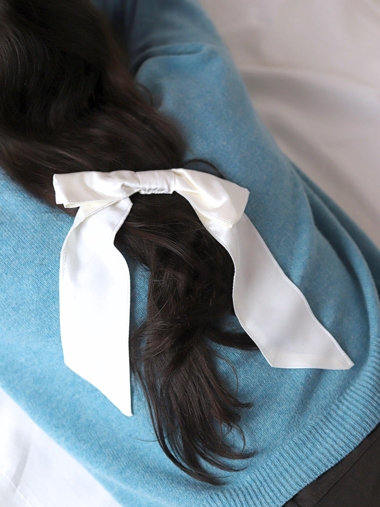 person lying on their front with brown hair wears a large ivory bamboo silk satin hair bow clip. they wear a red blue knit jumper on a plain background