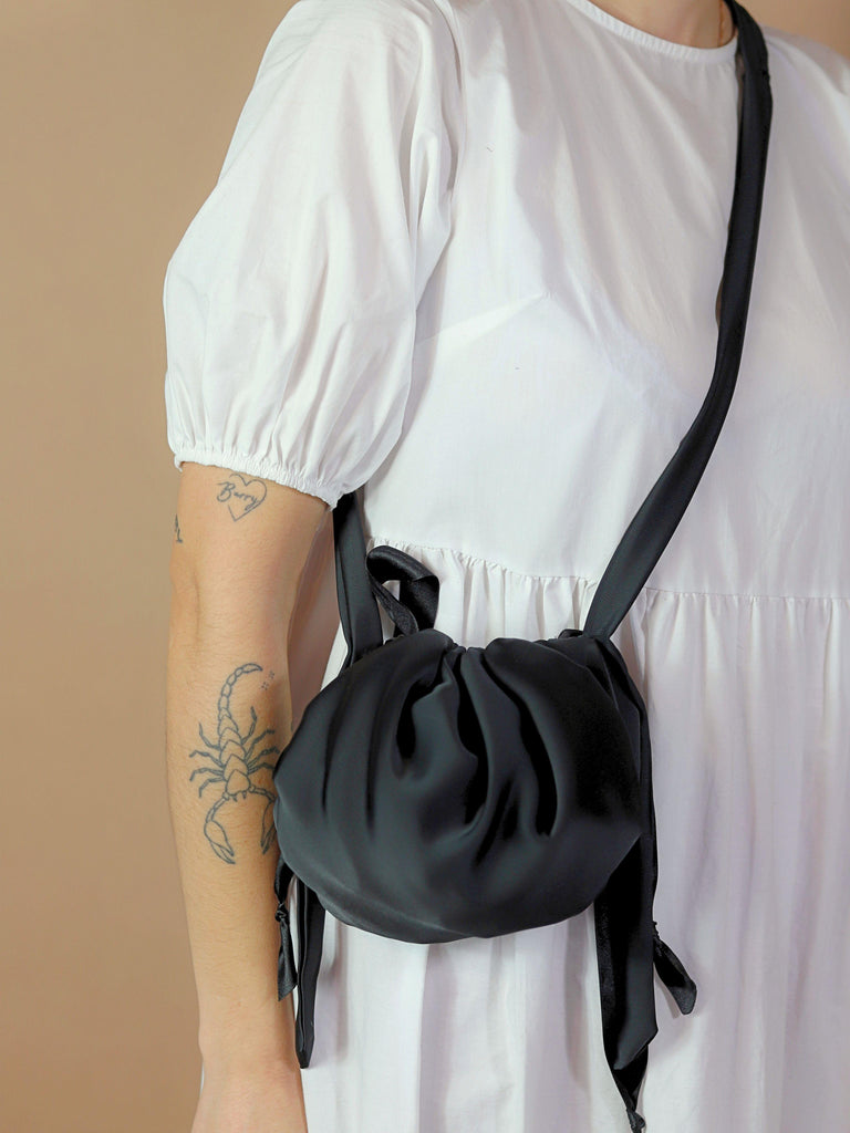 Person wearing a white dress and a crossbody black satin puffball drawstring bag, set against a beige background.
