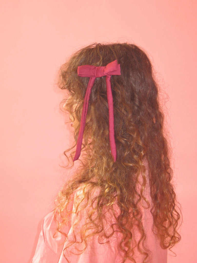Person with brown curly hair wearing a burgundy satin bow hair clip and a pink top against a pink background