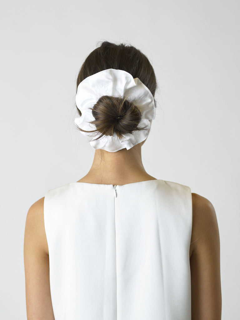 Person wearing a white sleeveless dress facing away wears an ivory bamboo silk satin scrunchie over their bun, set on a white background.