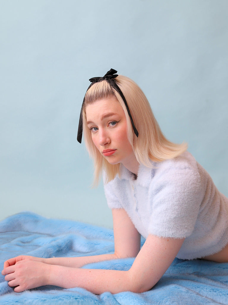Person with a black satin bow in their hair sitting on a blue faux fur blanket against a light blue background