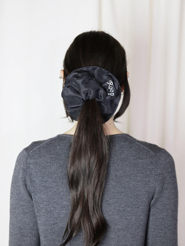 Person with long dark hair tied in a ponytail with a black scrunchie against a white background, they're wearing a grey knit cardigan.