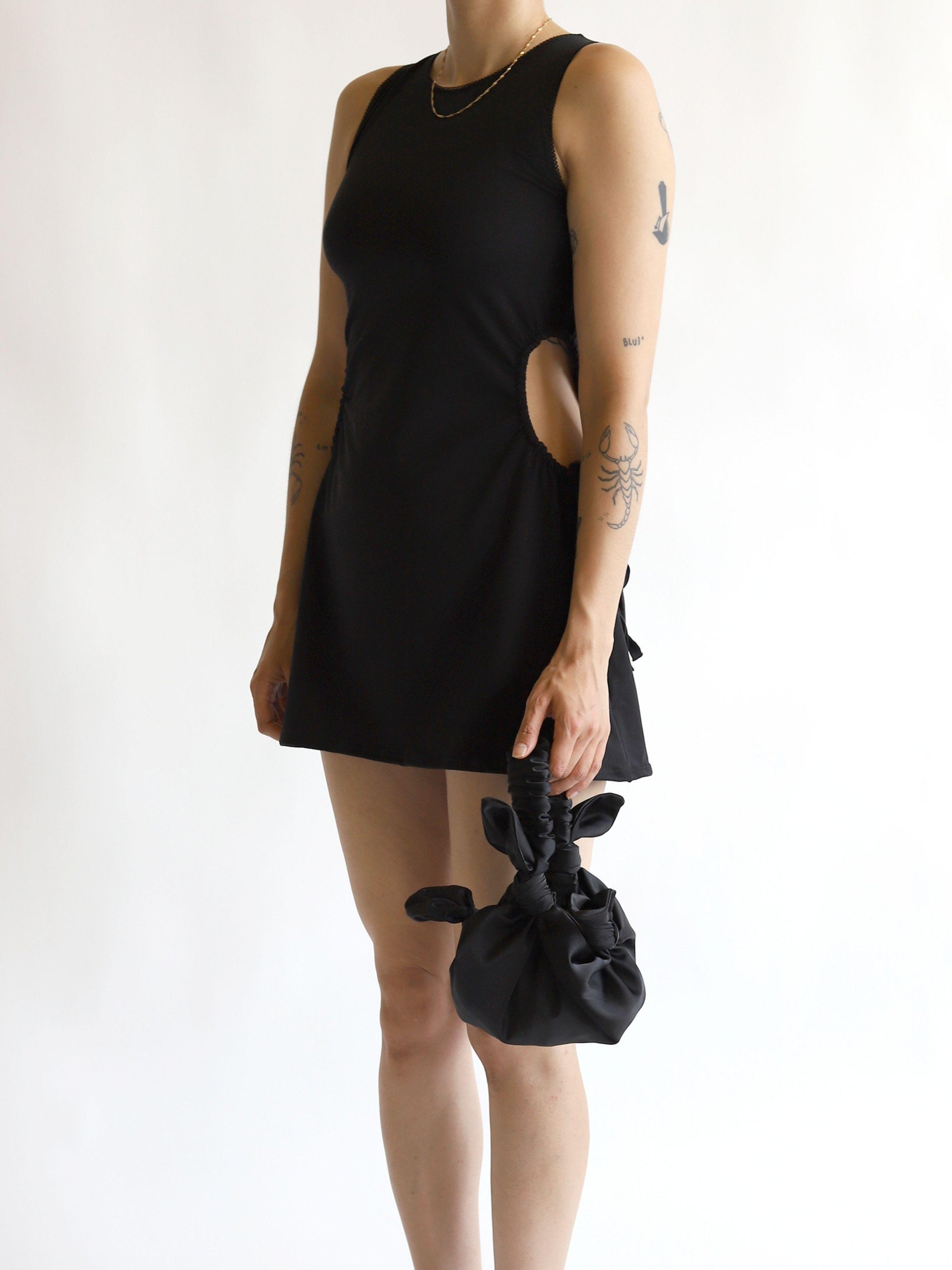 Person wearing a black sleeveless dress holding a black satin knotted bag on a white background