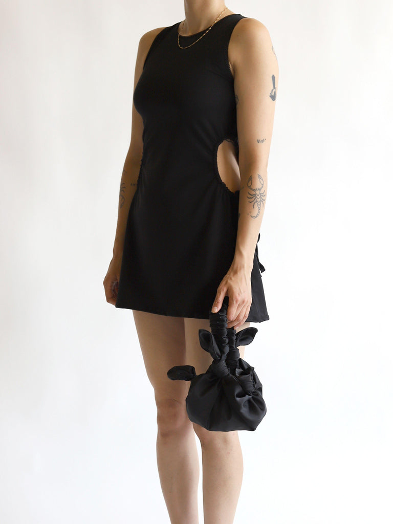 Person wearing a black sleeveless dress holding a black satin knotted bag on a white background