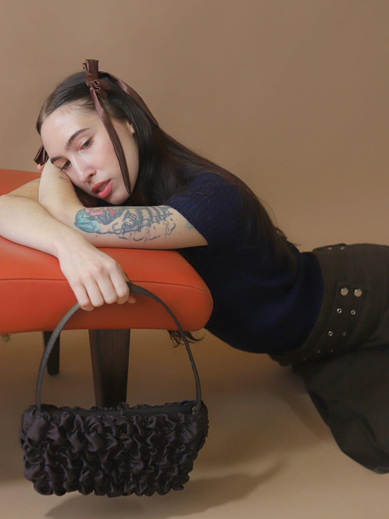 Person with tattoos wearing a blue ribbed jumper and a brown skirt, holds a brown textured handbag. They are leaning on a brunt orange chair, set on a beige background.