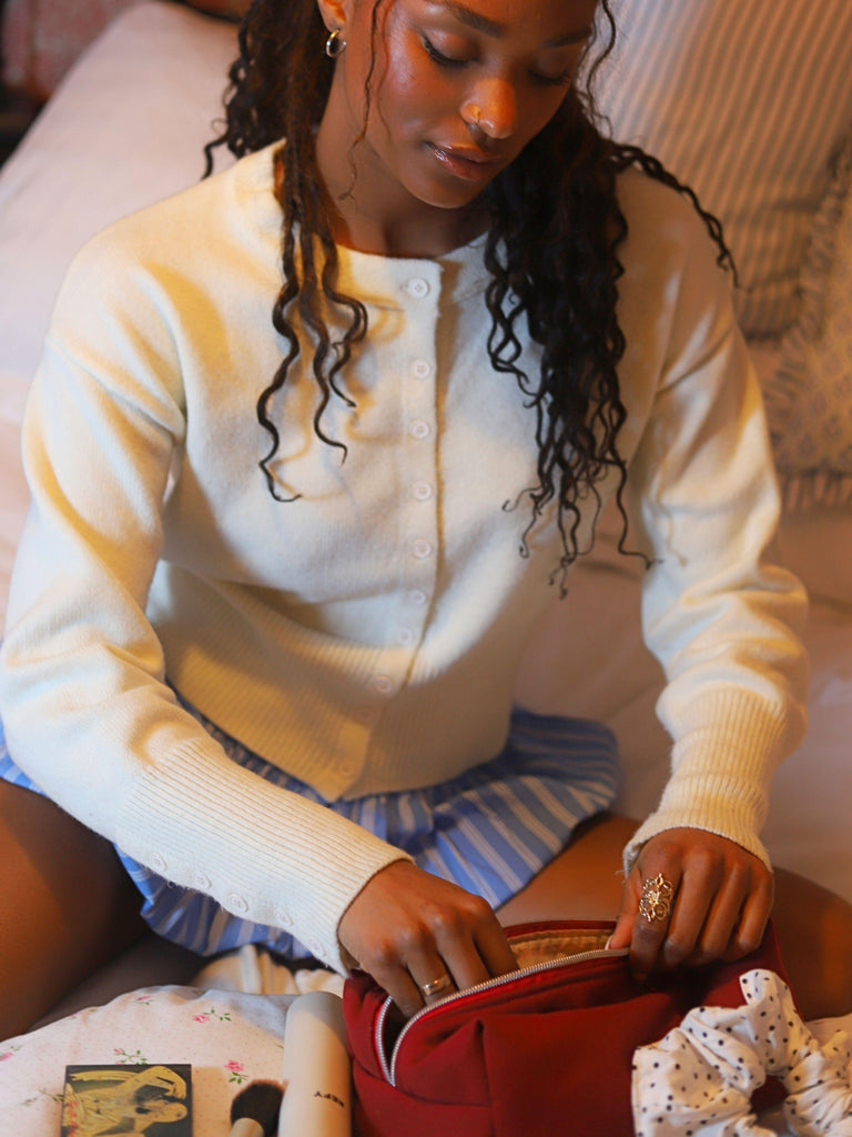 Woman in a white cardigan and striped blue shorts sits on a bed and looks inside her burgundy toiletry bag.