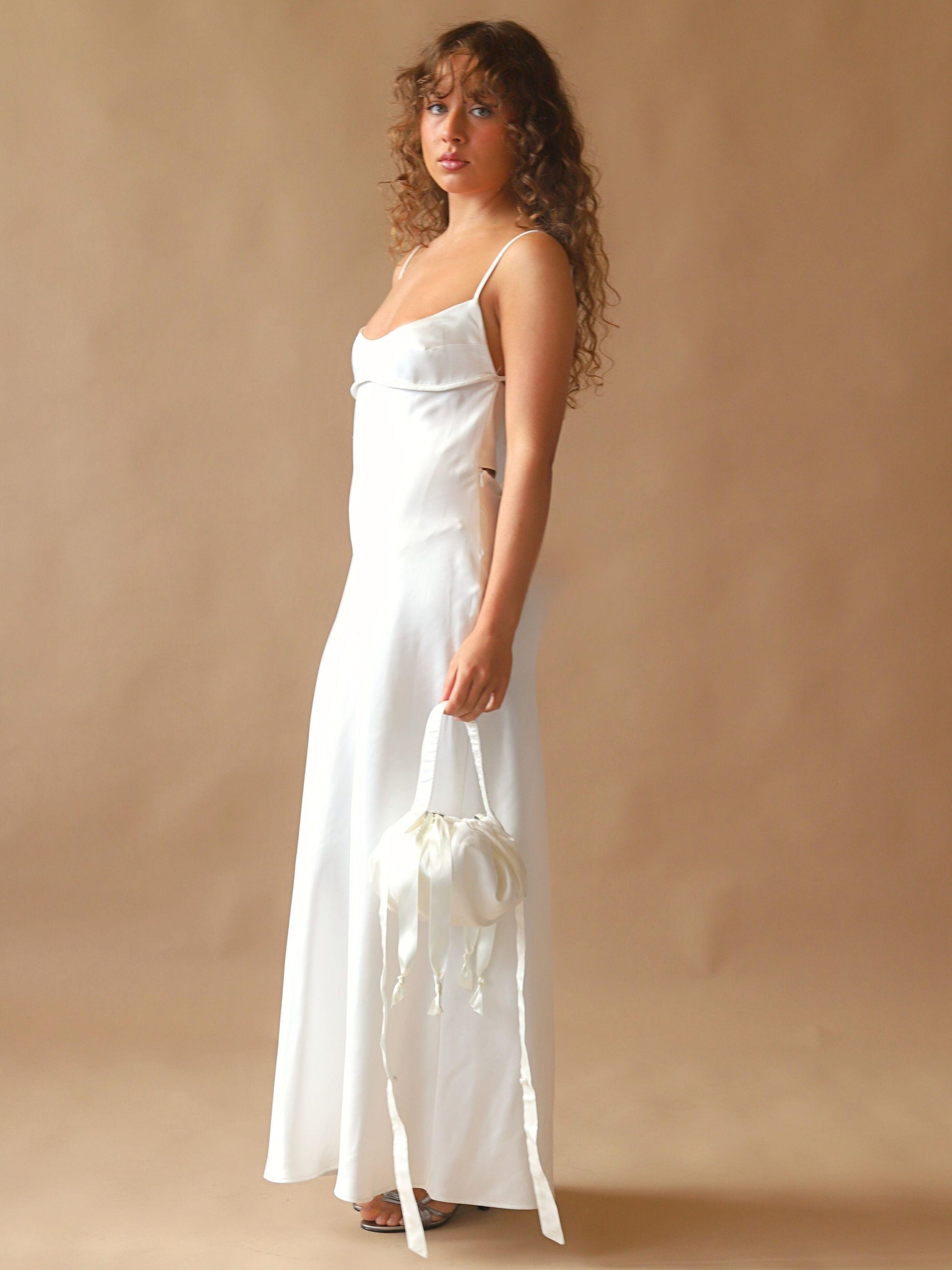 Person wearing an ivory satin slip dress, holds an ivory bamboo silk satin puffball drawstring handbag, set against a beige background.