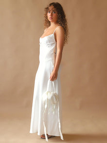 Person wearing an ivory satin slip dress, holds an ivory bamboo silk satin puffball drawstring handbag, set against a beige background.