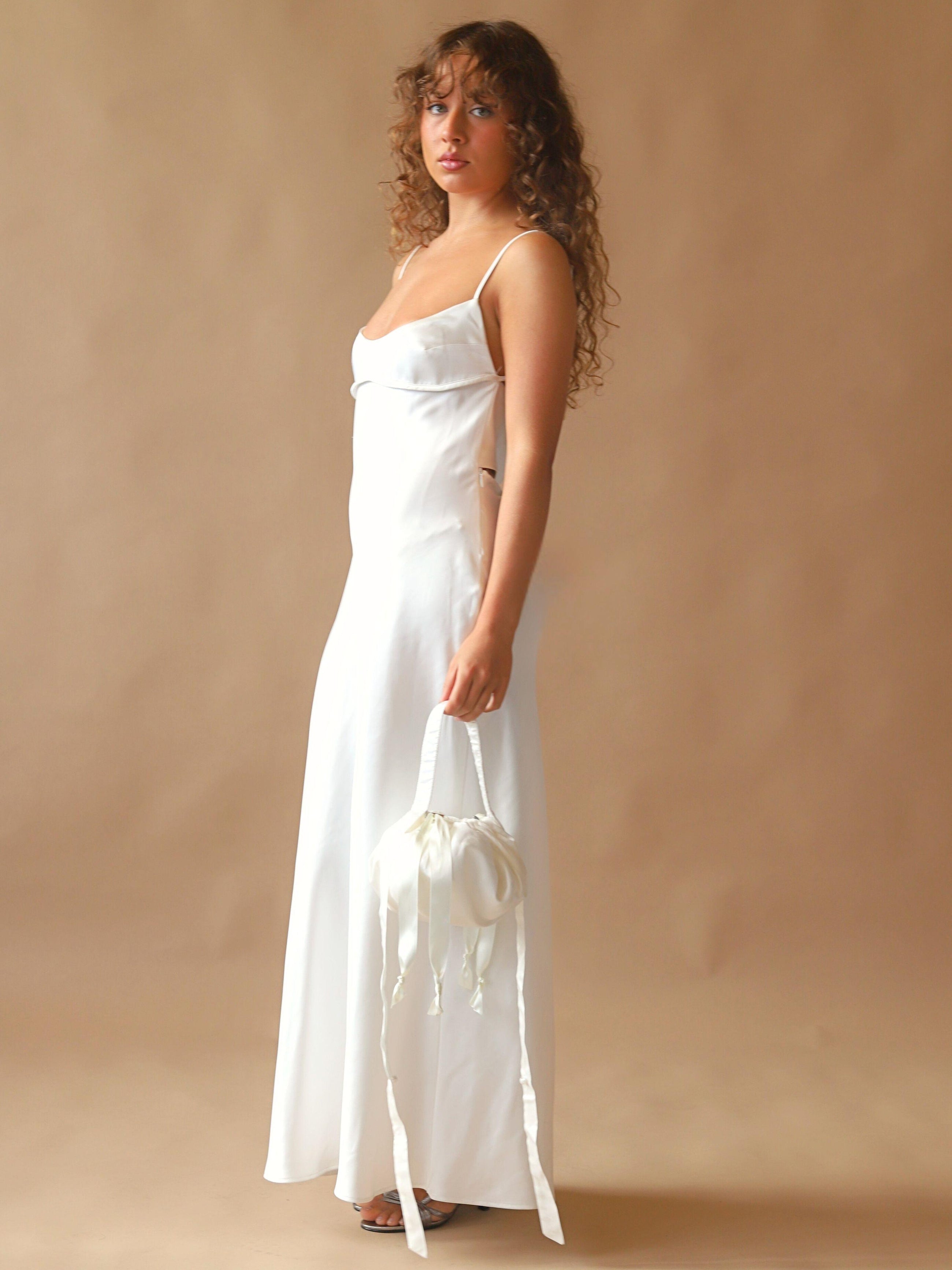 Person wearing an ivory satin slip dress, holds an ivory bamboo silk satin puffball drawstring handbag, set against a beige background.