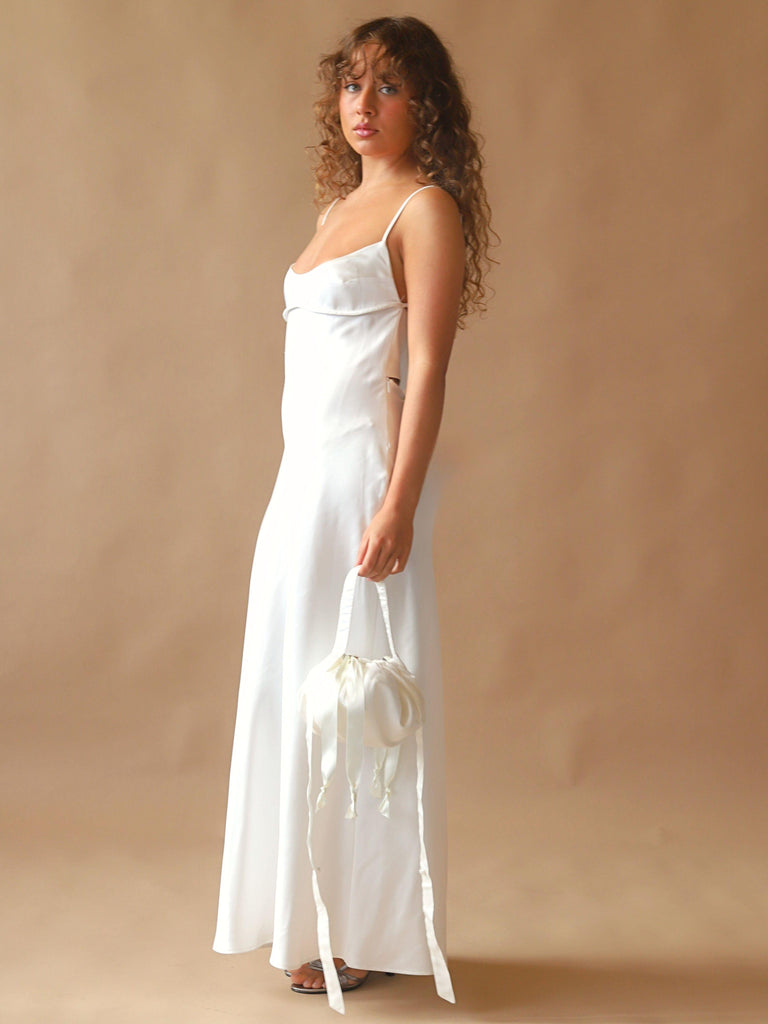 Person wearing an ivory satin slip dress, holds an ivory bamboo silk satin puffball drawstring handbag, set against a beige background.