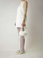 Person wearing a short ivory wedding dress, sheer white stocking and heeled ivory ballet pumps, holds an ivory bamboo silk satin puffball drawstring handbag, set against a plain background.