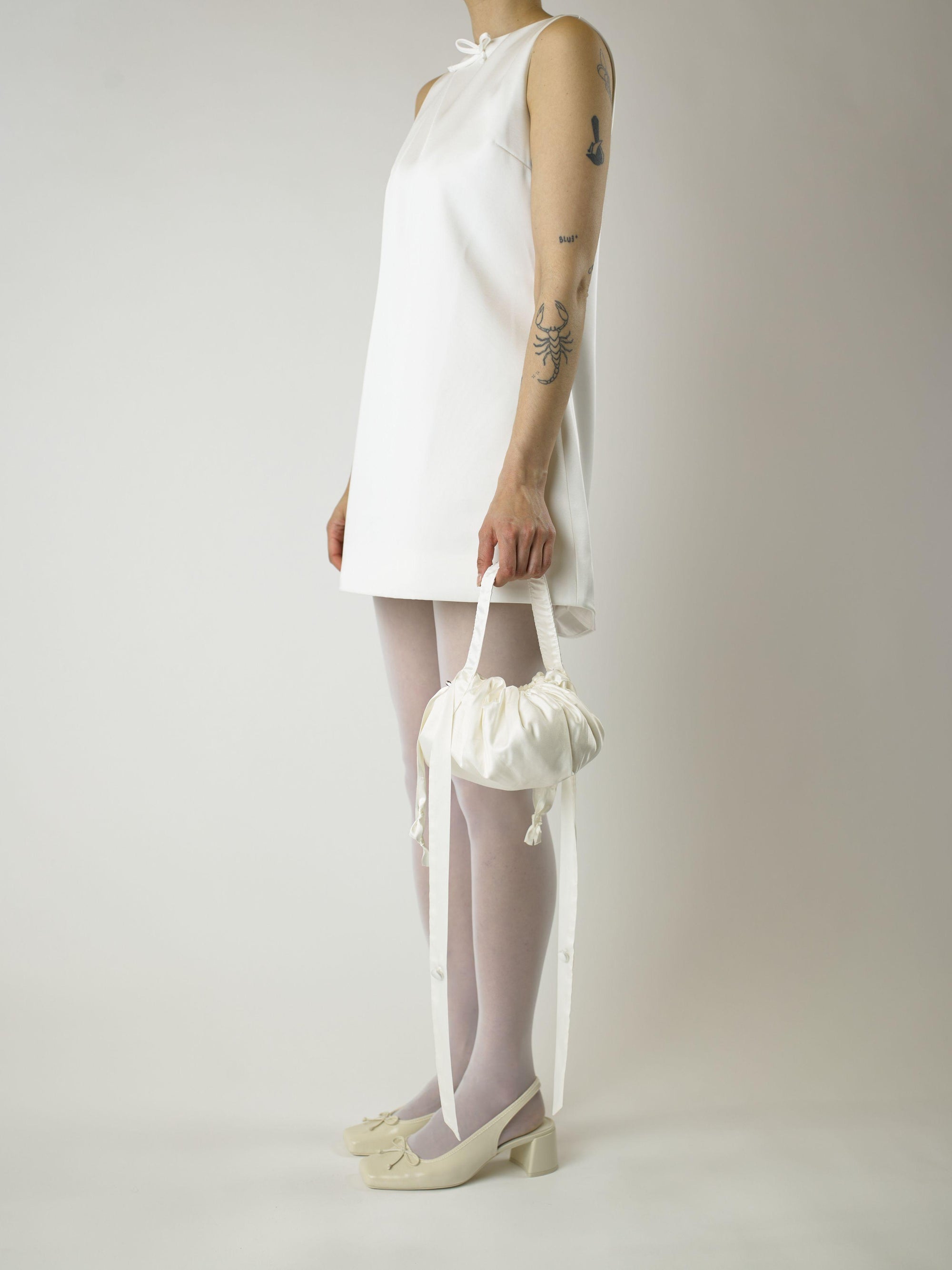 Person wearing a short ivory wedding dress, sheer white stocking and heeled ivory ballet pumps, holds an ivory bamboo silk satin puffball drawstring handbag, set against a plain background.