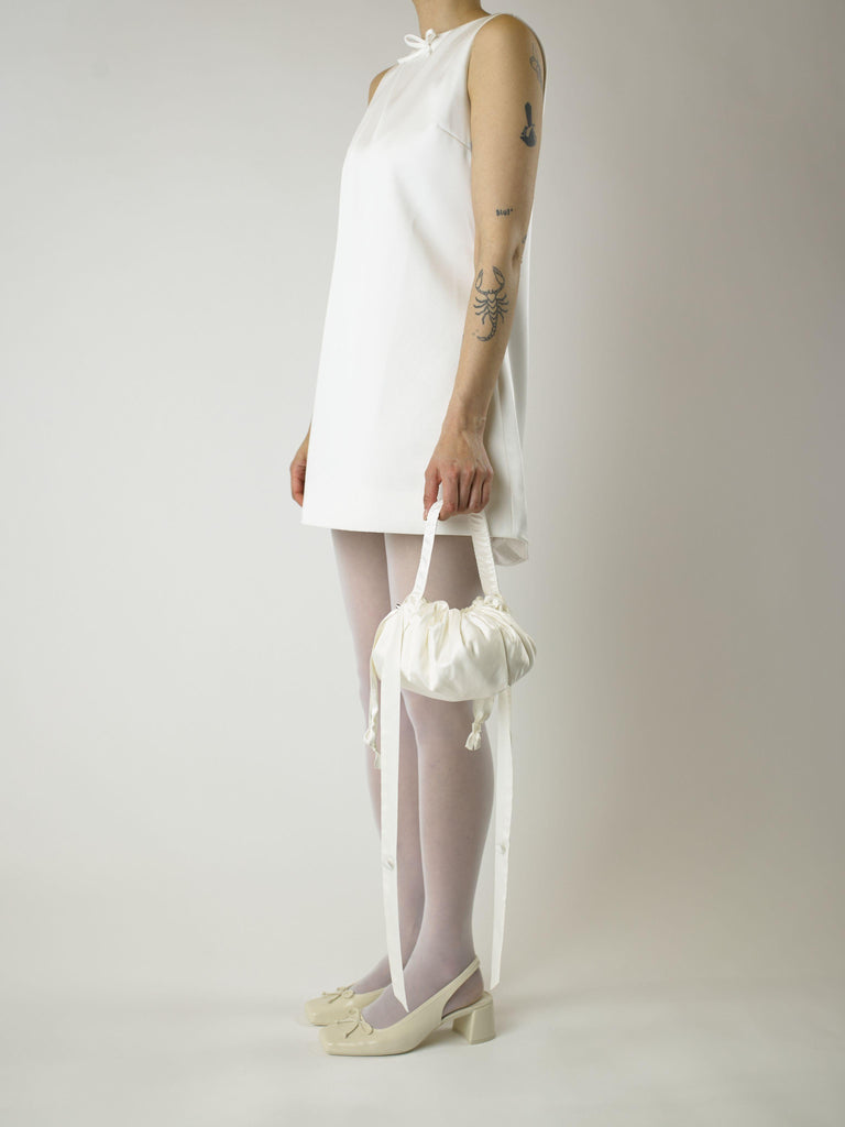 Person wearing a short ivory wedding dress, sheer white stocking and heeled ivory ballet pumps, holds an ivory bamboo silk satin puffball drawstring handbag, set against a plain background.