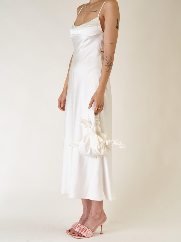 person wears an ivory satin slip dress with a pink ruched heels. They hold a ivory bamboo silk satin knotted handbag against a plain background