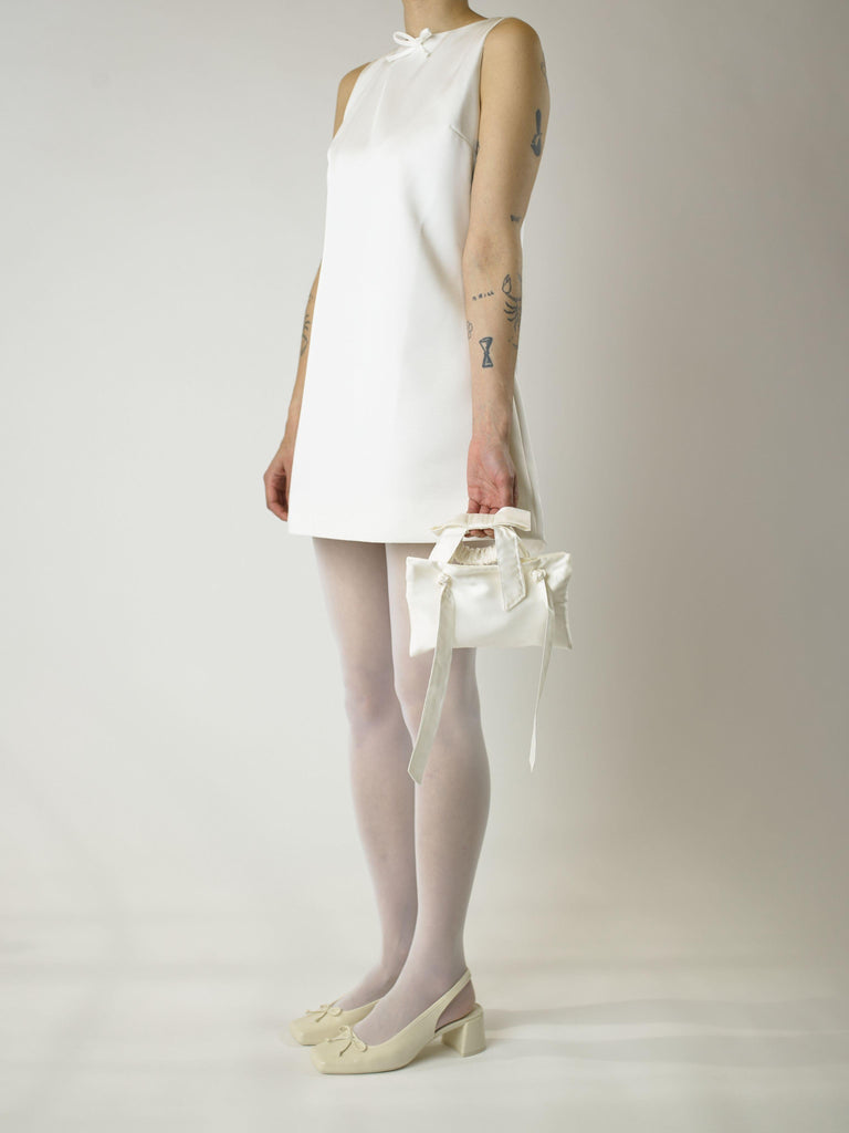 Person wearing a short ivory sleeveless wedding dress with sheer white stockings and heeled ivory ballet shoes. They hold an ivory bamboo silk satin wristlet bracelet bridal. Set on plain background