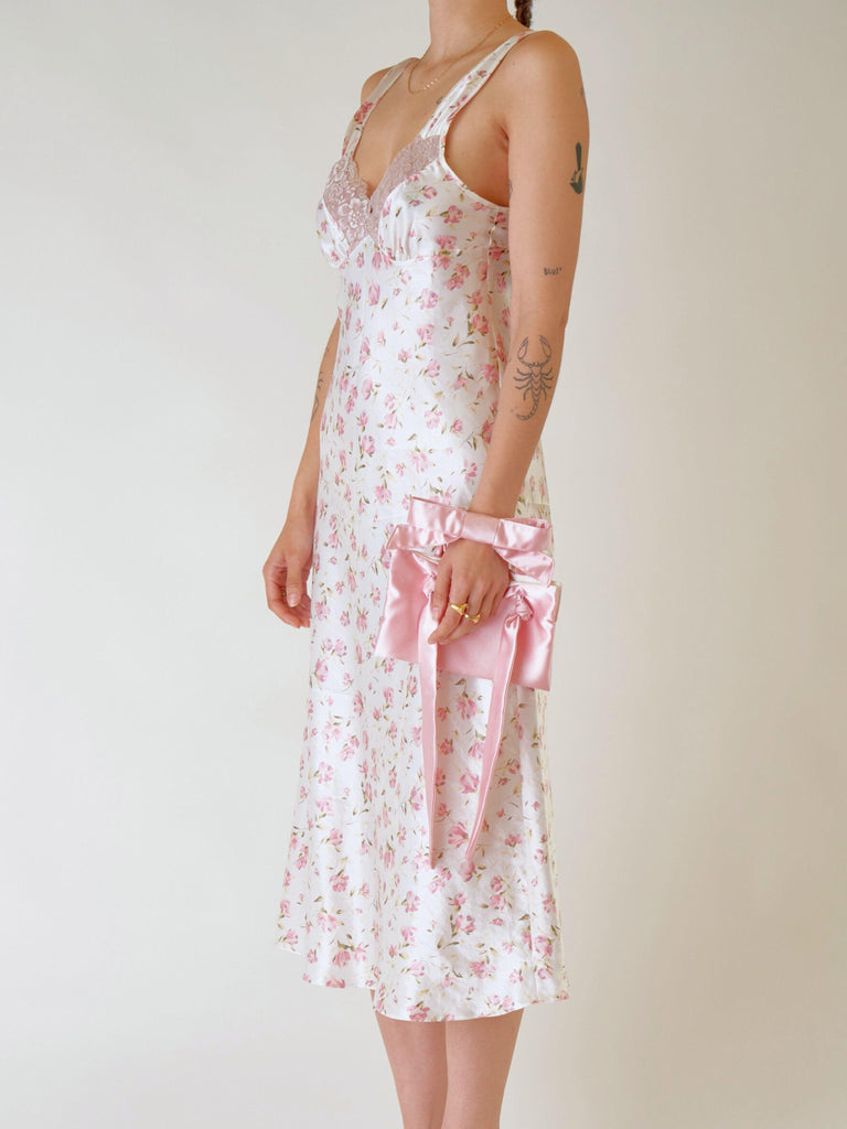 Woman wearing a floral dress with a pink satin bracelet bag on her wrist. Set on a plain background