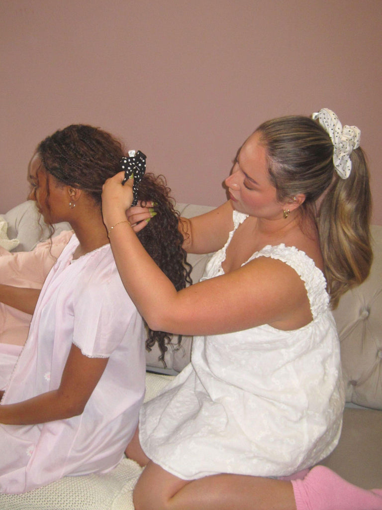 Two women in pyjama dresses sitting on a couch, one styling the other's hair. They are both wearing polka dot scrunchies.