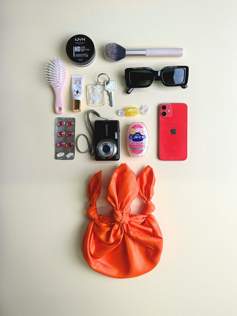Collection of everyday items including a phone, camera, and hairbrush on a light background. A orange handbag sits at the bottom.