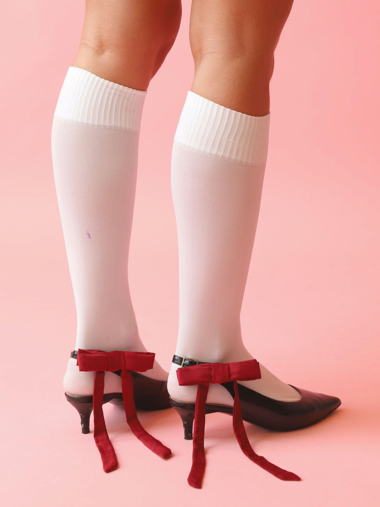 Person wearing black kitten heels and white knee-high socks with burgundy satin bow hair clips clipped on them. Set on a pink background