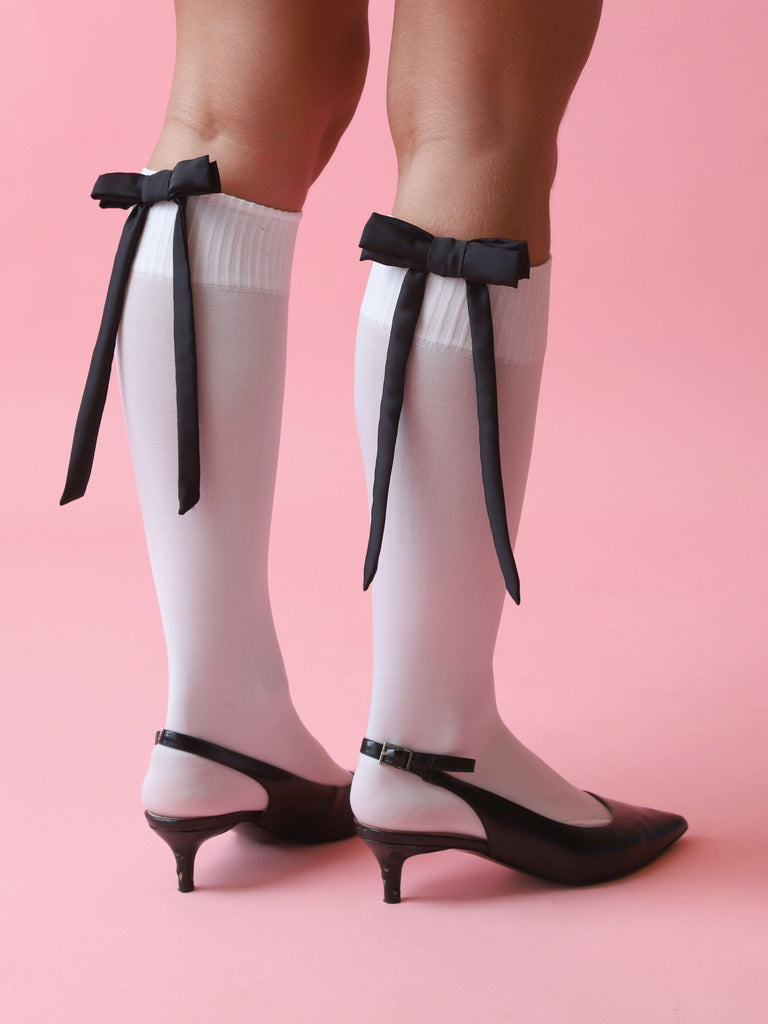 White knee-high socks styled with black bow hair clips and black kitten heels on a pink background