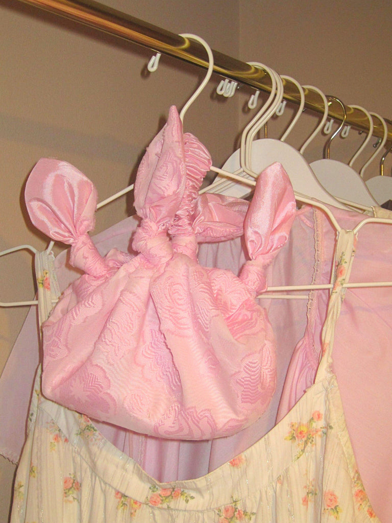 pink floral chiffon knotted handbag hangs on a white hanger  on a gold clothes rail with floral and pink garments behind it. Beige painted walls.