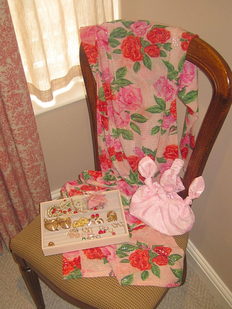 Floral sequinned dress draped over a wooden chair, an o[en jewellery tray and pink knotted handbag sits on the chair. Beige walls and a red patterned curtain and a sheer voile curtain cross the window.
