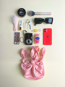 Collection of personal items including a pink satin bag, phone, camera, and makeup products on a light background. Pink knotted handbag sits at the bottom.