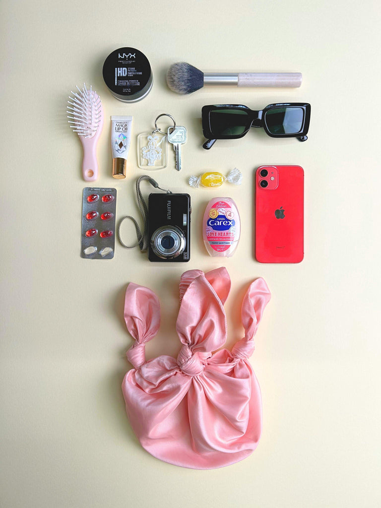 Collection of personal items including a pink satin bag, phone, camera, and makeup products on a light background. Pink knotted handbag sits at the bottom.