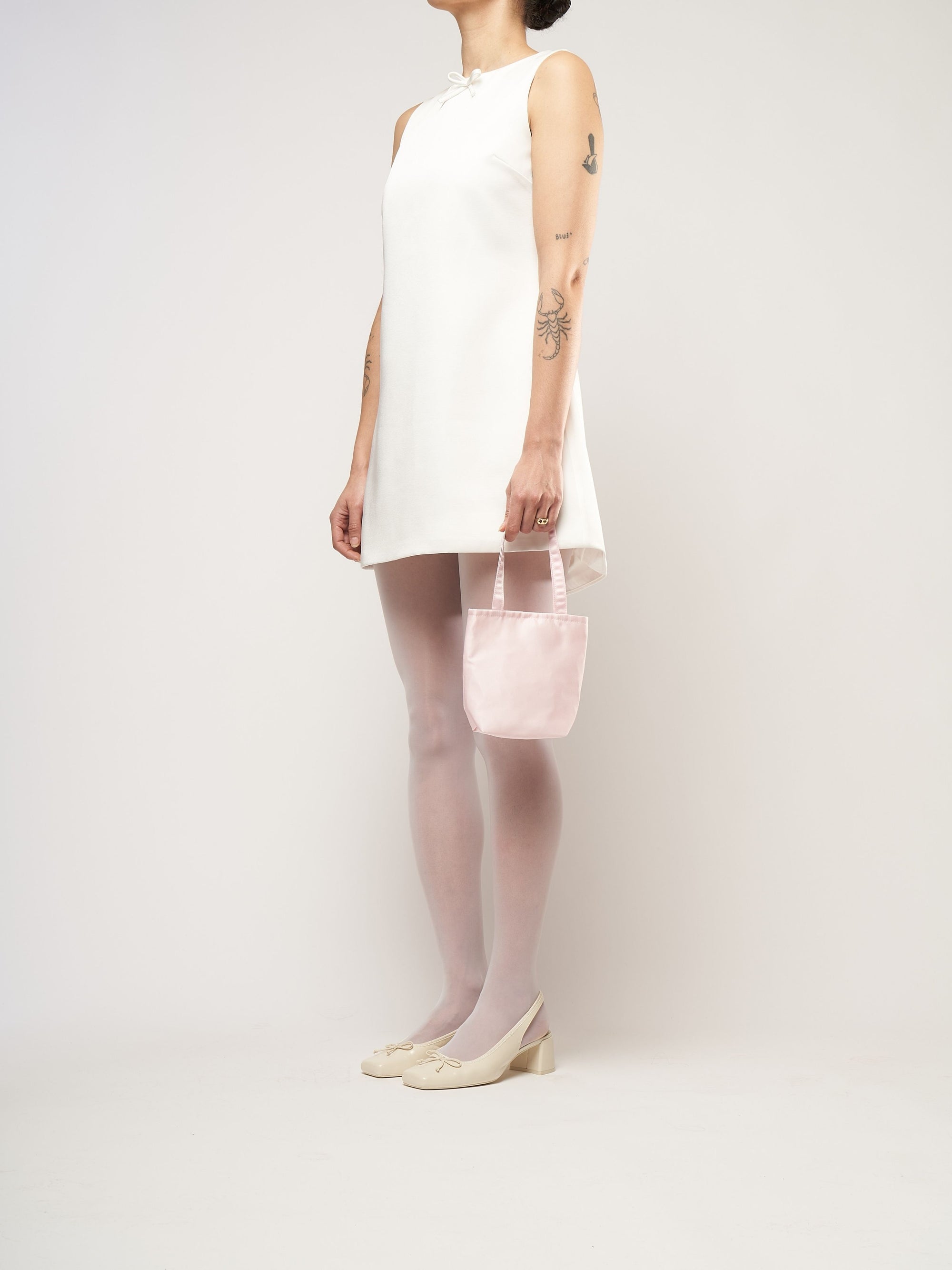 A person in a white sleeveless dress stands against a plain background. They hold a small light pink bag and wear white tights with cream heels.