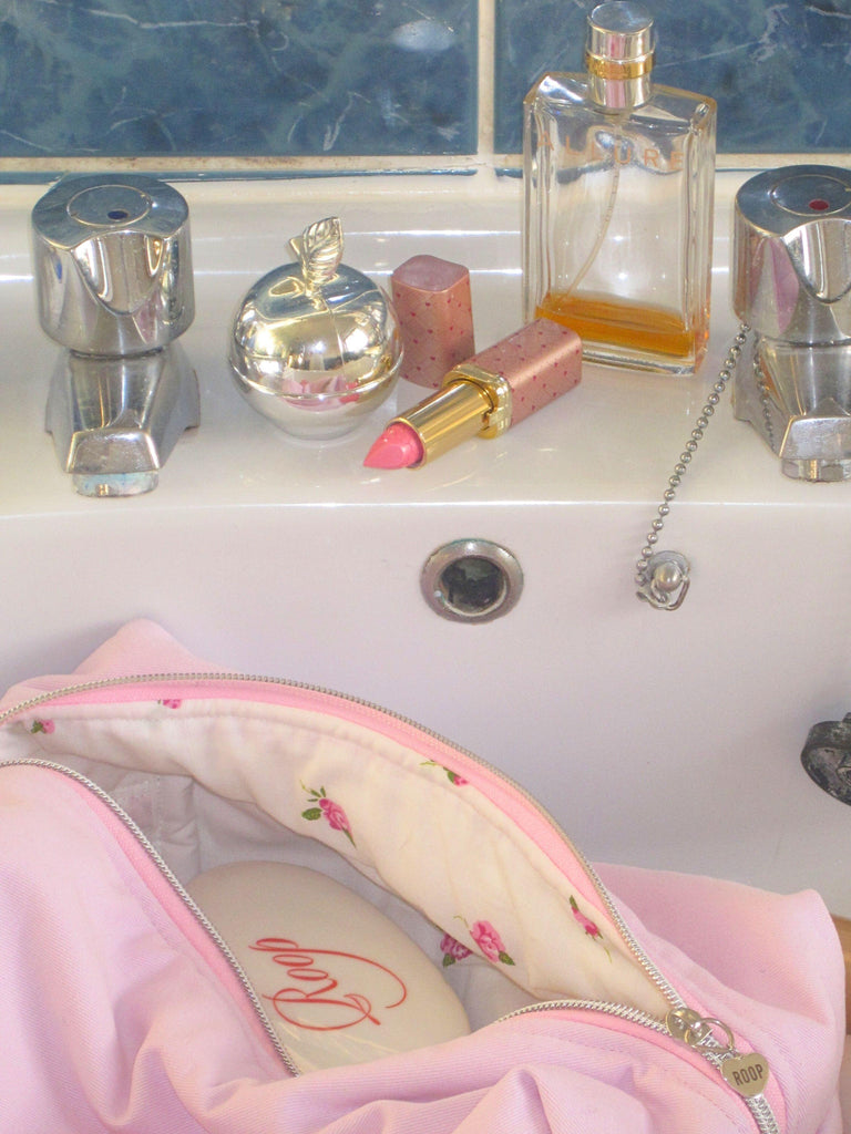 Pink makeup bag with floral design in a bathroom sink with lipsticks and perfumes.