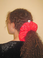 Person with curly brown hair wears a large pink scrunchie in a ponytail, they have a black velvet top on and silver hoop earrings, set against on beige background