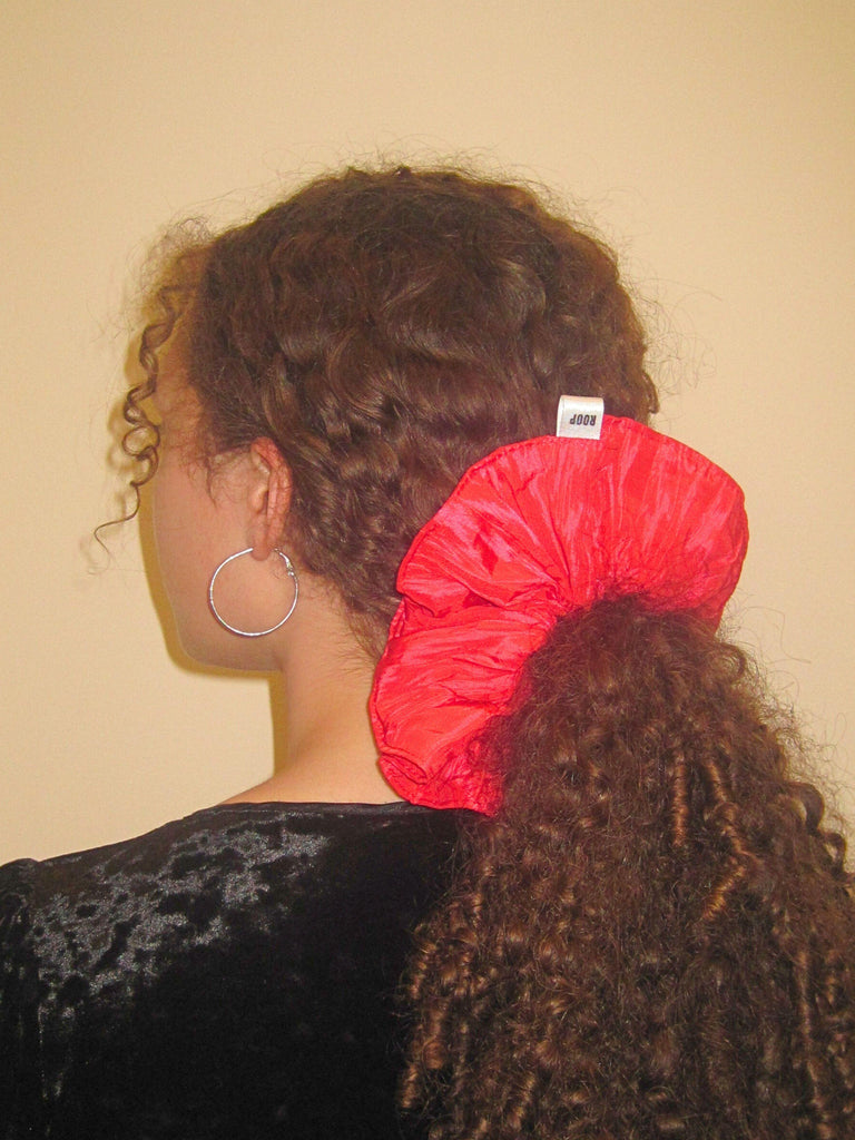 Person with curly brown hair wears a large pink scrunchie in a ponytail, they have a black velvet top on and silver hoop earrings, set against on beige background