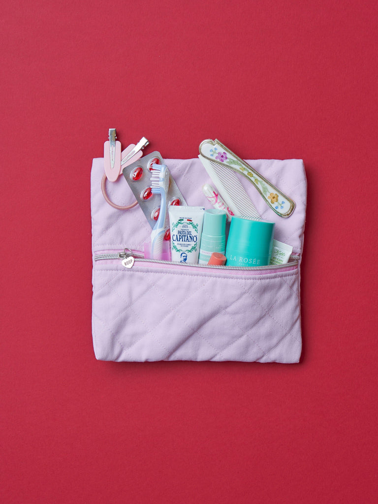 A quilted pink cotton zipper pouch, the zip has a heart charm on it. Various are items are placed inside the open zip including a toothbrush, floral hair comb and deodorant. Set on a red background.