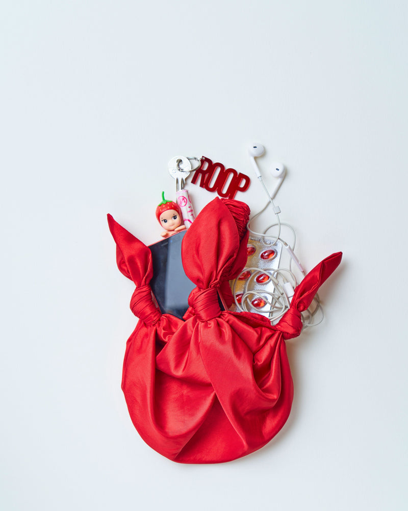 A red fabric bag holds a smartphone, a toy figure, earbuds, and a red keychain with "ROOP" text. The bag's playful knot design adds a whimsical touch.
