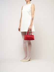 A woman in a sleeveless white dress holds a red handbag. She wears white tights, cream heels, and has tattoos on her arms, standing against a plain background.
