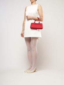 A woman in a sleeveless white dress holds a red handbag. She wears white tights, cream heels, and has tattoos on her arms, standing against a plain background.