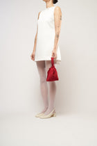 A person in a white sleeveless dress stands against a plain background. They hold a small red bag and wear white tights with cream heels.