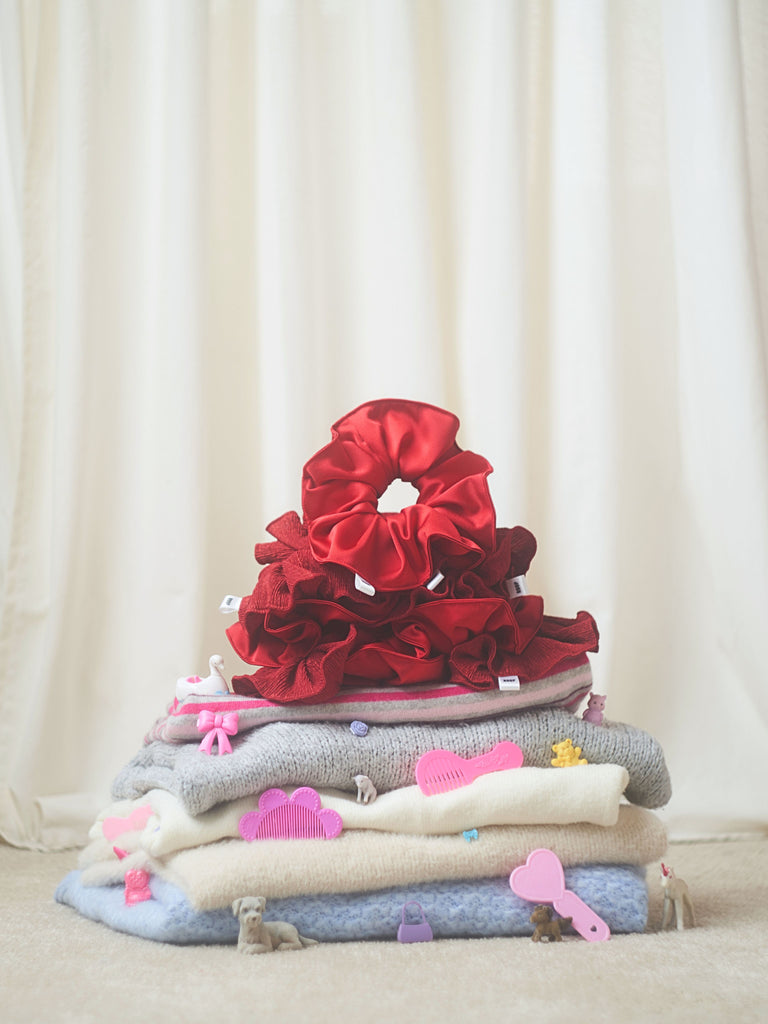 Stack of folded knitted jumpers with red scrunchies on top of them. Set against a cream curtain background