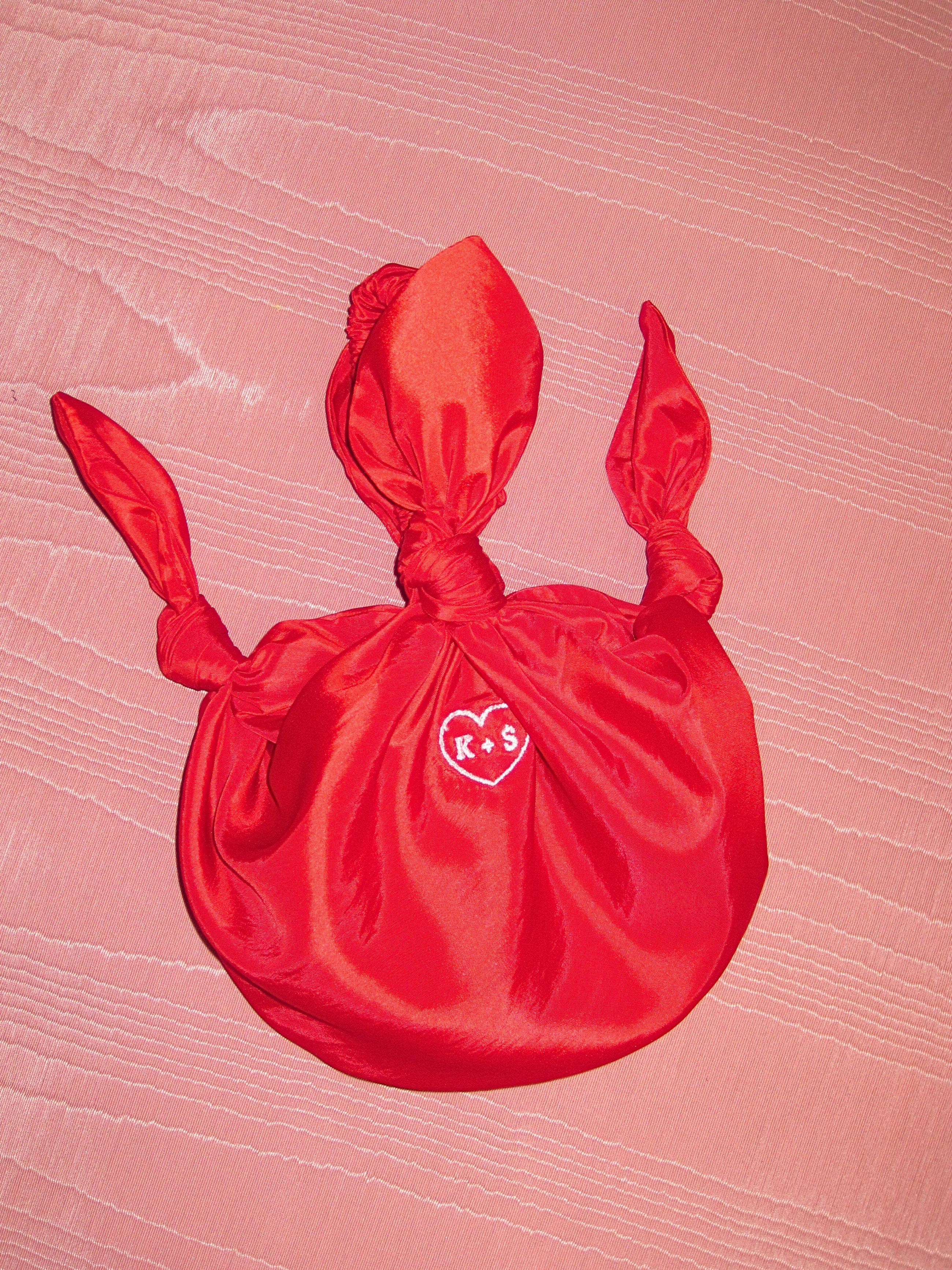 red taffeta baby furoshiki handbag with white embroidery of a heart with K+S in the centre. Set on pink moire fabric background.