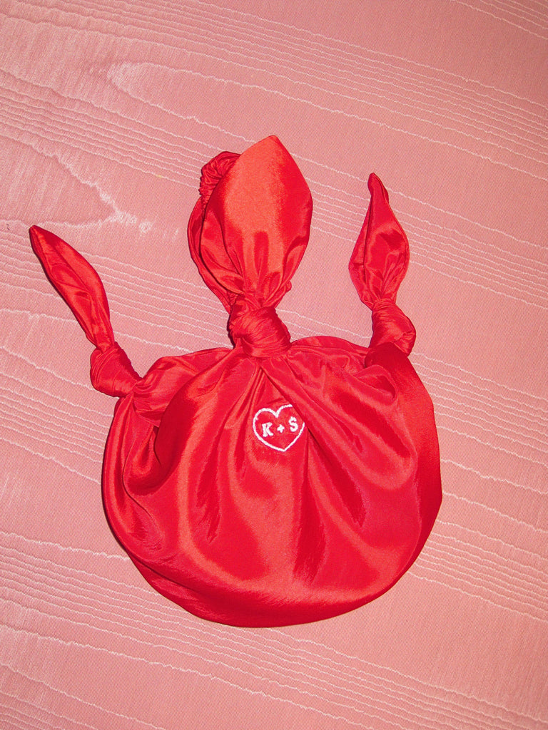 red taffeta baby furoshiki handbag with white embroidery of a heart with K+S in the centre. Set on pink moire fabric background.