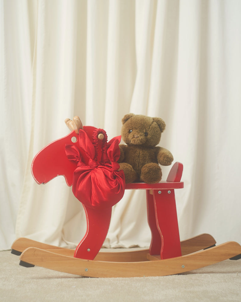 A brown teddy bear and a red knotted taffeta handbag sit on a red wooden rocking horse against a soft cream curtain backdrop, evoking a cozy, nostalgic feel.